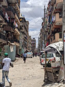 An unpaved road in one of Nairobi's neighborhoods, with mid-rises lining the streets; residents dry their laundry from balconies. Cars and pedestrians traverse the street, without sidewalks.