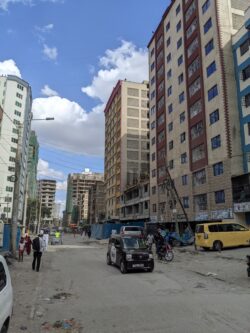 Another Nairobi neighborhood with newer high rises. The road is still unpaved, and pedestrians traverse narrow paths between the buildings and the street.