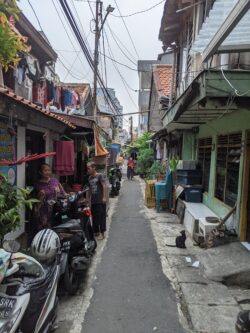 Two buildings flank a narrow alley in Jakarta. Clothes hang from one and motorcycles are parked to the left, leaving a narrow passageway.